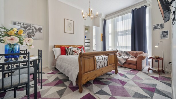 Une chambre à coucher avec un lit, un banc en bois, un bureau et une chaise. Il y a une fenêtre avec des rideaux, un lustre et une lampe.