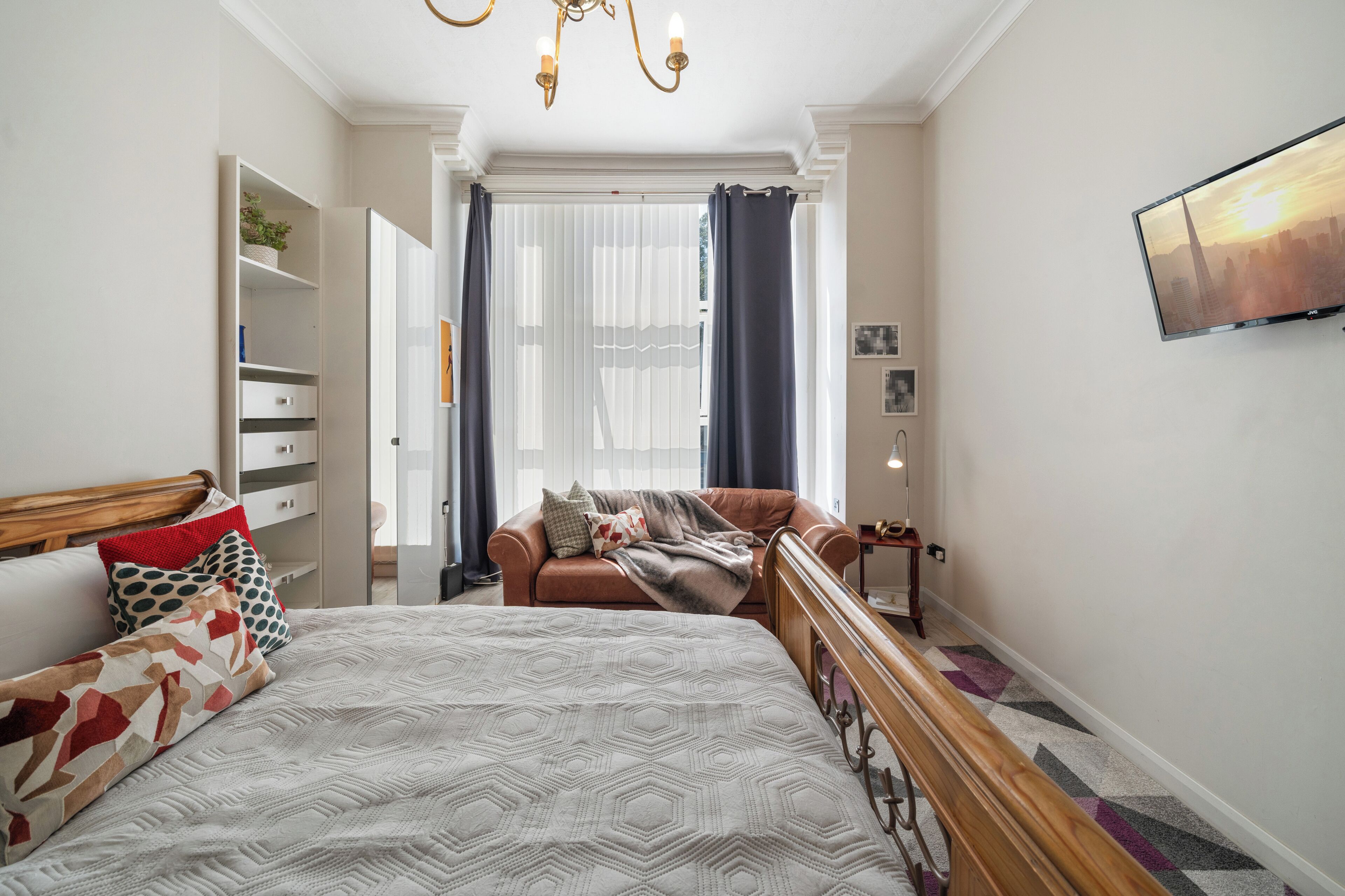 Une chambre avec un grand lit, une tête de lit en bois, un canapé en cuir marron, un téléviseur fixé au mur et une fenêtre ornée de rideaux légers.