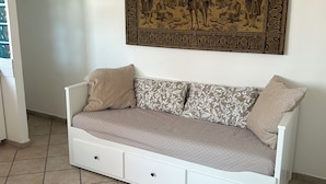 Un canapé blanc agrémenté de coussins à motifs, une tapisserie décorative au-dessus, et un sol carrelé.