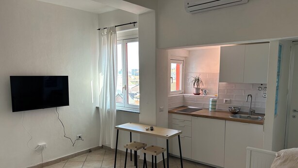 Un espace de vie compact comprenant un téléviseur fixé au mur, une petite table à manger avec des tabourets, un coin cuisine avec un évier et un plan de travail, et un radiateur.