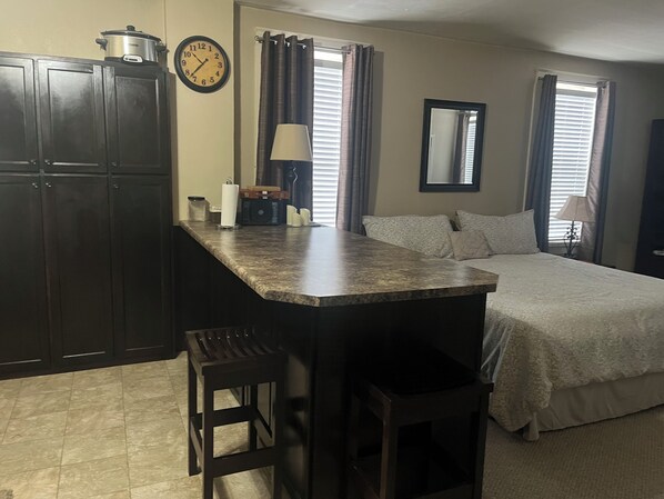 Un dormitorio pequeño con cama, una kitchenette con estufa, un reloj y una ventana con cortinas.