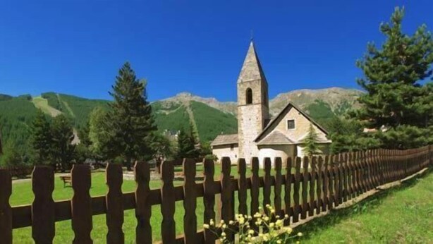 A church with a tall bell tower and a small adjacent building, surrounded by a wooden fence and greenery. - Hotel Las Donnas (Saint-Etienne-de-Tinee)