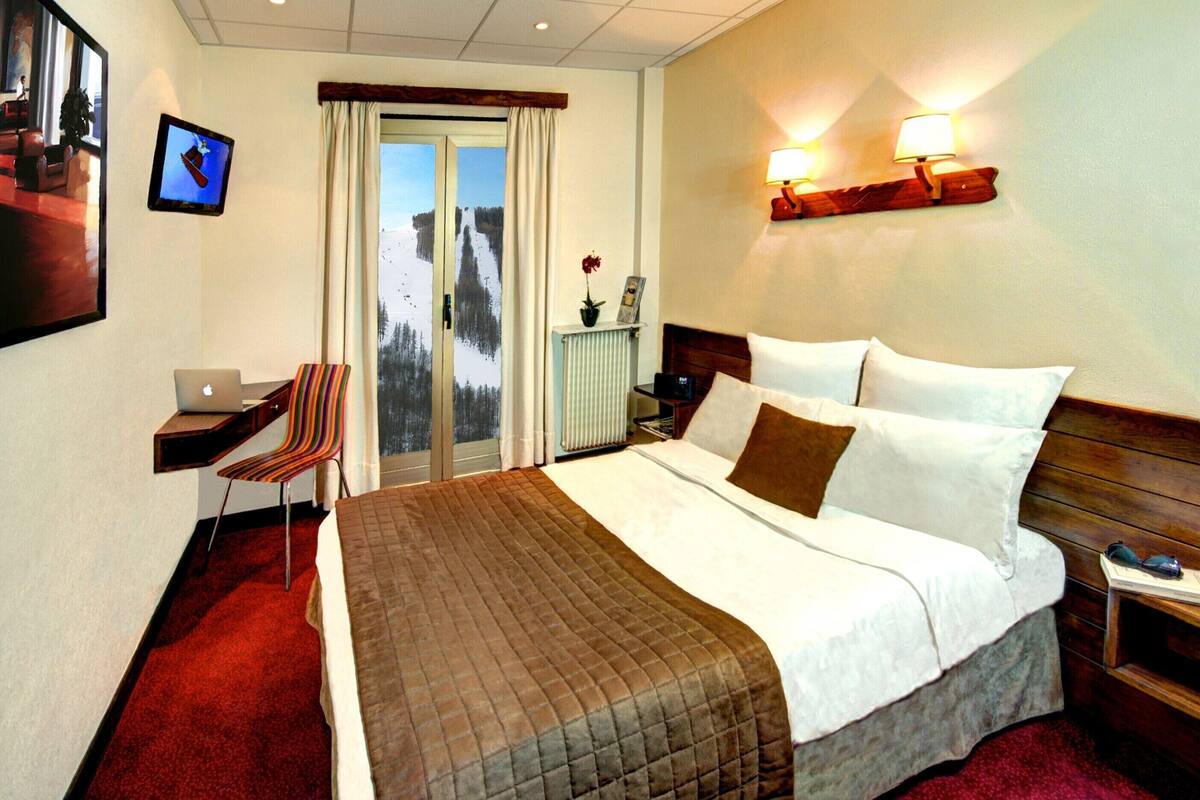 A hotel room with a bed, a desk with a laptop, a chair, a window with a view of a snowy landscape, and a wall-mounted lamp.