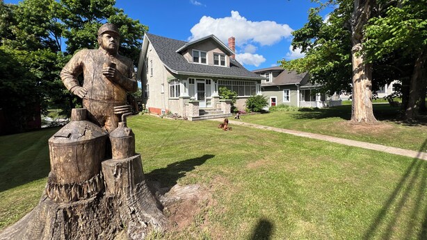Una escultura de madera de un hombre con sombrero y overol, una casa con porche y un césped bien cuidado.