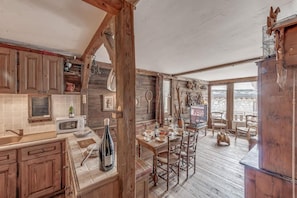 A rustic kitchen with wooden cabinets, a dining table, and chairs. There is a wooden ceiling and a view of the outside through large windows.
