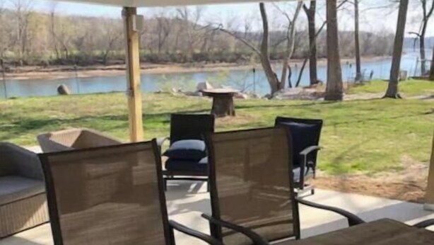 Patio esterno con tavolo e sedie in legno, zona relax coperta e vista su un lago.