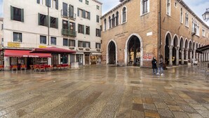 A spacious public square with stone flooring, a bar named "Bar Stellina" with outdoor seating, and a building with arched entrances.