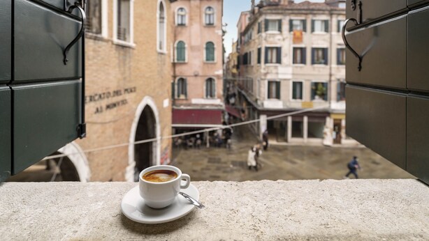 Una taza de café sobre una mesa con vista a una calle a través de una ventana abierta.