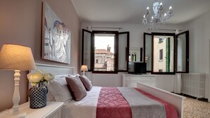 Ein Schlafzimmer mit einem Bett, einem Nachttisch, einer Lampe, einer Vase mit Blumen, einem Gemälde, einem Spiegel und Blick auf Gebäude vor dem Fenster.