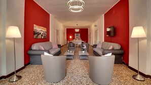 Eine Hotelhalle mit roter Akzentwand, zwei Sofas, einem Couchtisch, einem an der Wand befestigten Fernseher und einer Stehlampe.