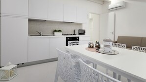 Moderni keittiö, jossa on valkoiset kaapit, valkoiset tuolit ruokapöydän ympärillä, mikroaaltouuni ja pieni kasvi työtasolla.