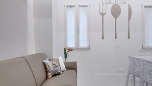 Moderni olohuone, jossa on beigenvärinen sohva, koristeellinen tyyny, pieni pöytä valkoisella pöytäliinalla ja seinä, joka on koristeltu keittiöaiheisella seinämaalauksella.