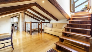 Ein geräumiges Loft mit freiliegenden Holzbalken, einer Holztreppe, einem beigen Sofa und Blick ins Freie.