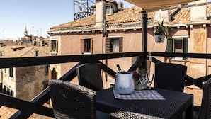 Eine Terrasse mit Korbmöbeln, einem Tisch mit einer Monetto-Tasse und Gläsern sowie Blick auf Gebäude mit Terrakotta-Dächern.