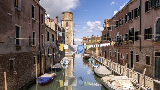 A narrow canal with boats docked on both sides, flanked by historic buildings with balconies and a tower in the background. - Arsenale Canal View 1 by Wonderful Italy (Venice)