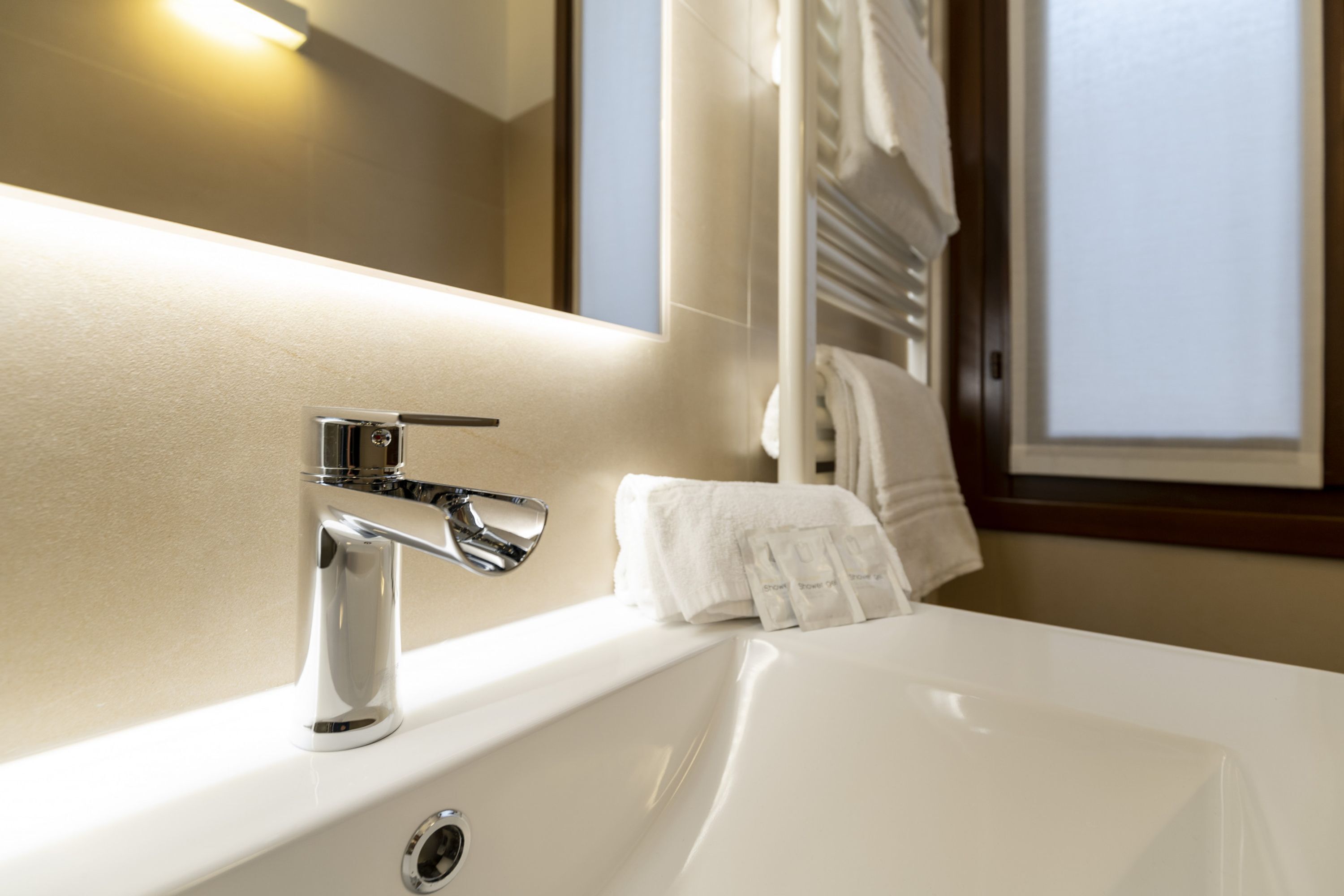 Ein modernes Badezimmer mit einem chromfarbenen Wasserhahn, einem weißen Waschbecken und einem Spiegel, der ein Fenster mit Vorhängen widerspiegelt.