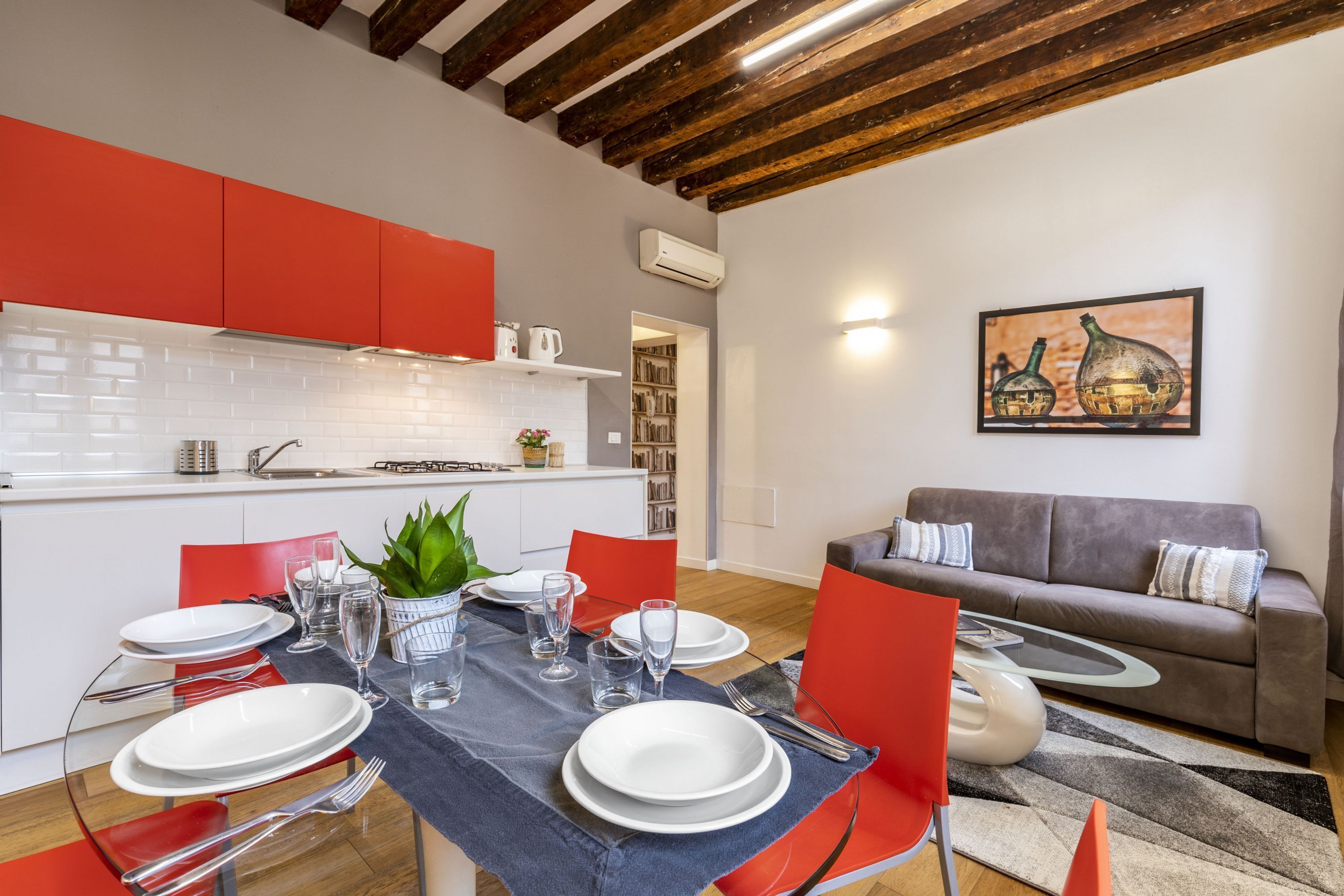 Una cocina moderna con armarios rojos, un comedor con una mesa dispuesta para cuatro personas, y una sala de estar con un sofá y un cuadro en la pared.