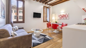 Een moderne woonkamer met een bank, een eethoek met rode stoelen, een keukeneiland en een aan de muur bevestigde televisie.
