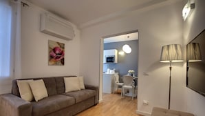 Una sala de estar moderna con un sofá marrón, una mesita y una lámpara. Se ve un área de cocina a través de un vano abierto.