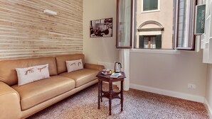 A cozy living room with a brown leather sofa, a small wooden table with a book and a kettle, a framed picture on the wall, and a window with a view of a building. - Rugabella Rialto by Wonderful Italy (Venice)
