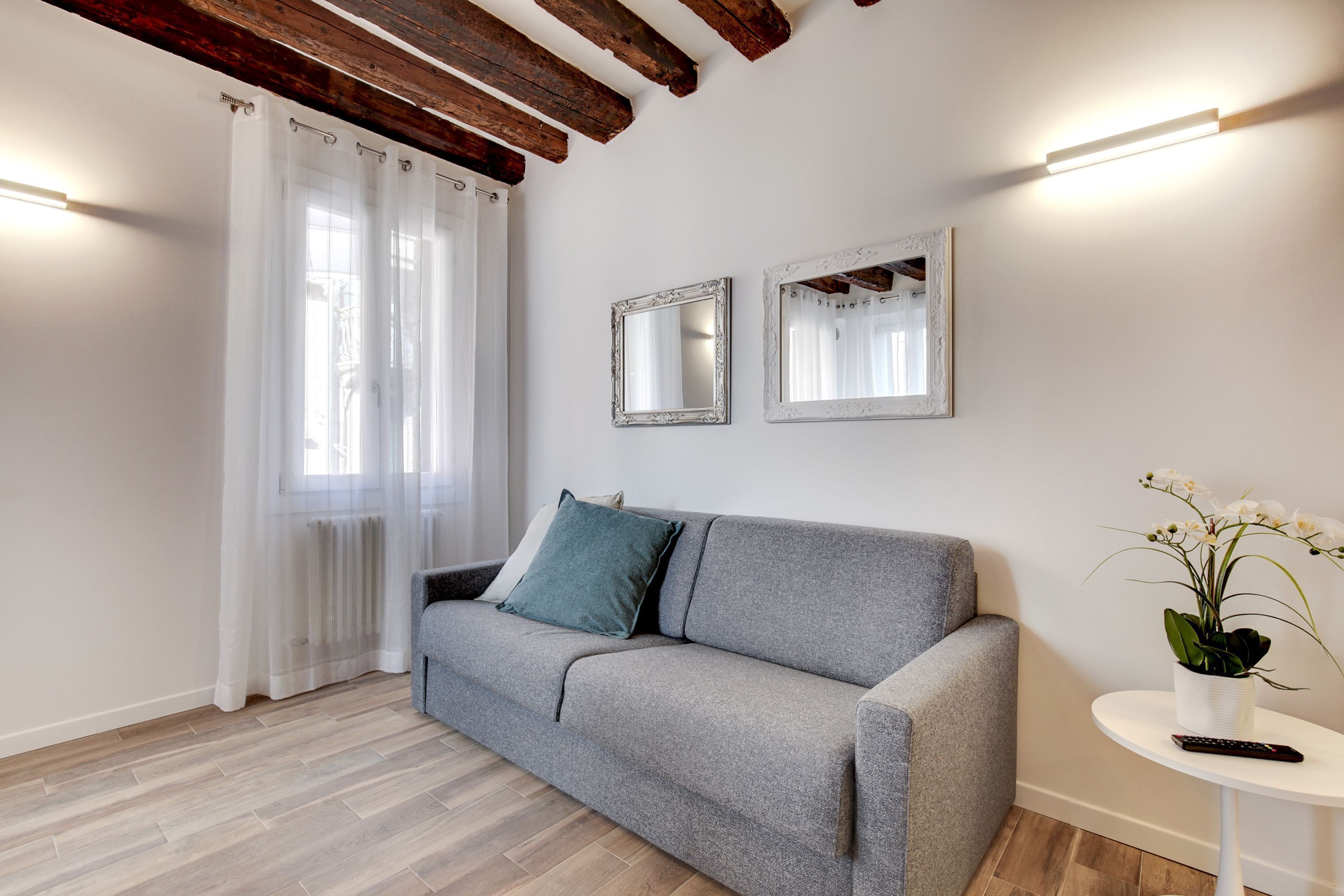 Un salon moderne avec un canapé gris, deux miroirs blancs, une petite table d’appoint blanche avec une télécommande et une orchidée en pot.