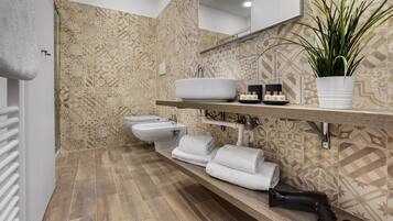 A modern bathroom with a large mirror, a white sink, a bidet, and a wooden shelf with towels and toiletries.
