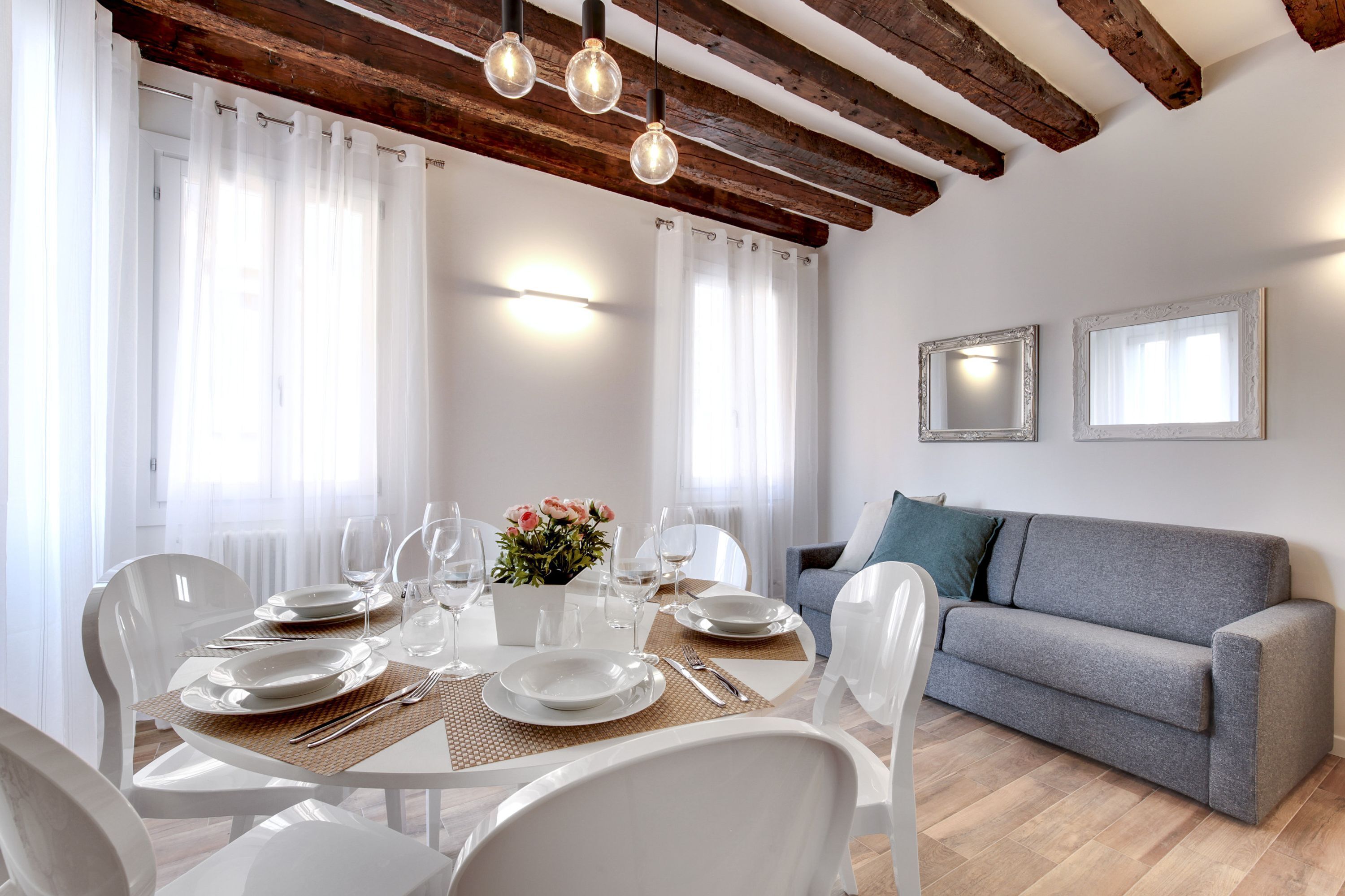 Un comedor con una mesa dispuesta para cuatro, sillas blancas y un sofá gris al fondo.