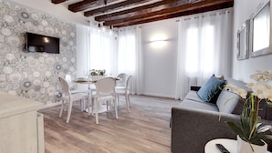 A modern living room with a dining area, wooden flooring, a flat-screen TV mounted on the wall, and a sofa with a potted plant. - Laguna Luxury 1 by Wonderful Italy (Venice)
