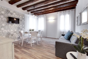 A modern living room with a dining area, wooden flooring, a flat-screen TV mounted on the wall, and a sofa with a potted plant. - Laguna Luxury 1 by Wonderful Italy (Venice)