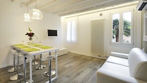 Un comedor moderno con una mesa blanca, sillas de vidrio transparente y un jarrón de flores. Hay un sofá blanco y un radiador cerca de una ventana con vistas a palmeras.