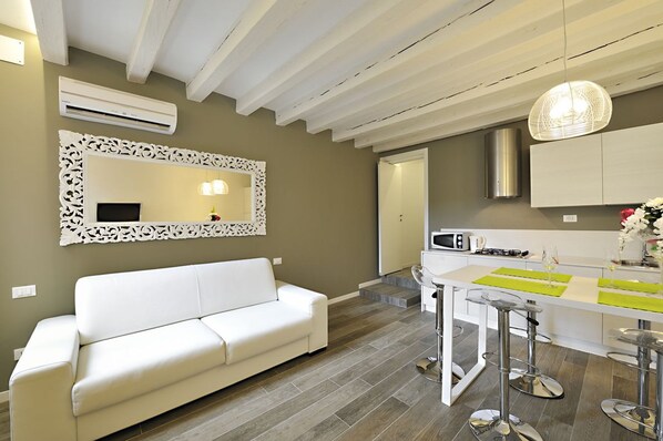 Una sala de estar moderna con un sofá blanco, una zona de cocina con microondas y una mesa de comedor con taburetes de bar.