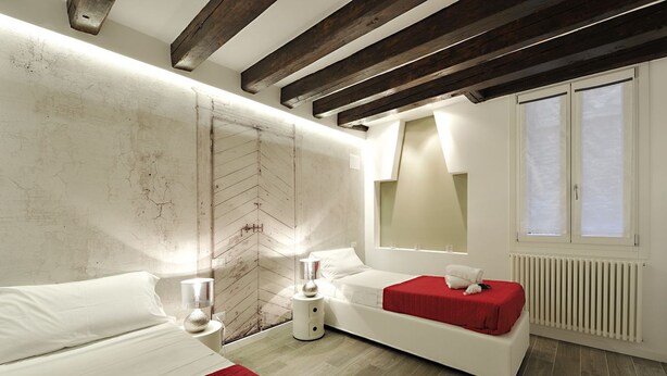 Un dormitorio moderno con una cama grande, un sofá cama más pequeño, una mesita de noche con lámpara, un espejo grande y una ventana con persianas.