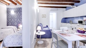 Une pièce compacte et bien éclairée, comprenant un lit, un canapé bleu, une table à manger avec des chaises et un coin cuisine.