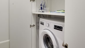 Une petite buanderie équipée d’un lave-linge à chargement frontal, d’étagères au-dessus pour le rangement et d’une porte dotée d’une poignée.