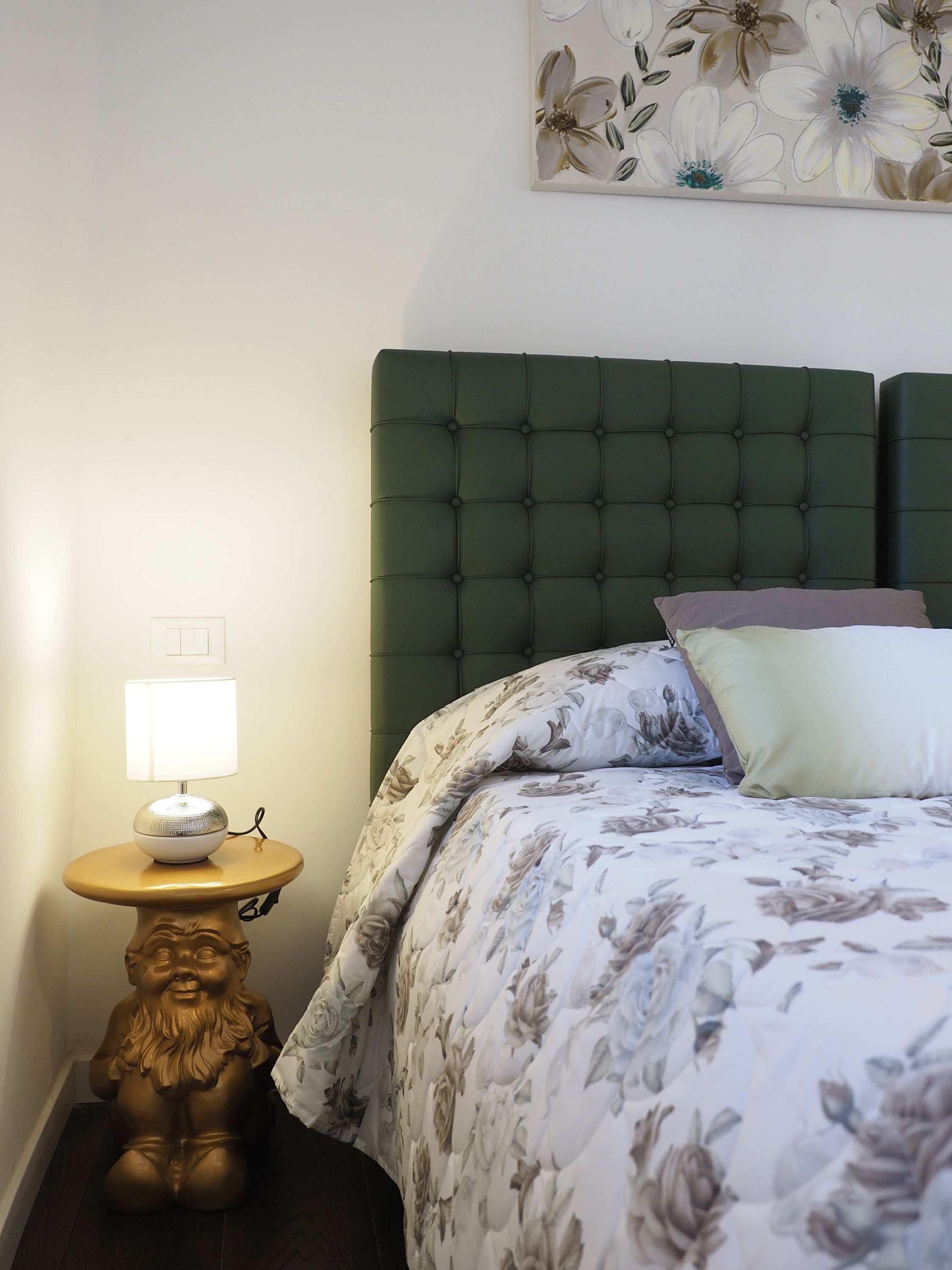 A bedroom with a green upholstered headboard, a floral-patterned bedspread, a bedside lamp, and a decorative lion statue.