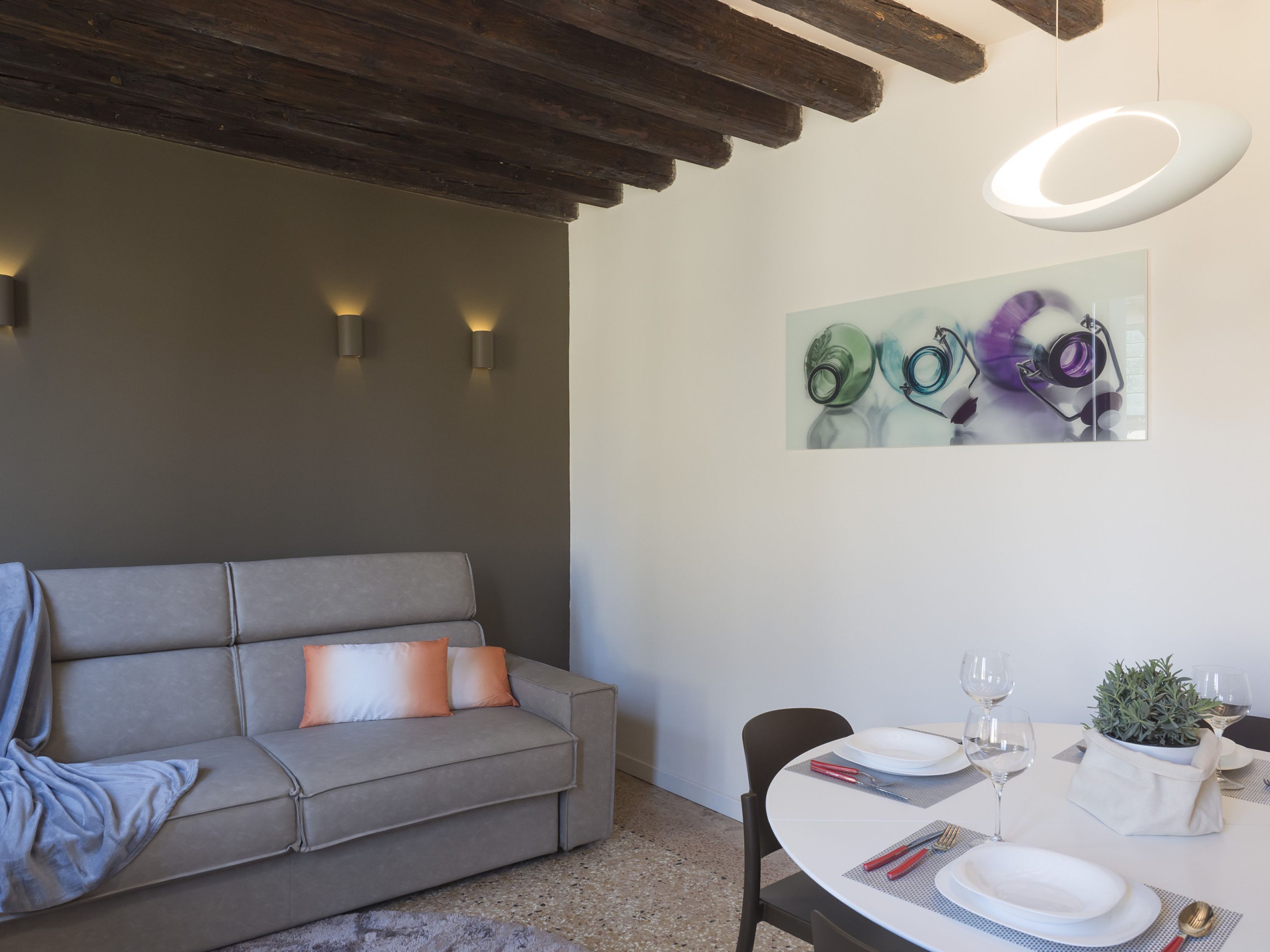 A modern living room with a grey sofa, a dining table set with plates, glasses, and cutlery, and a painting on the wall.