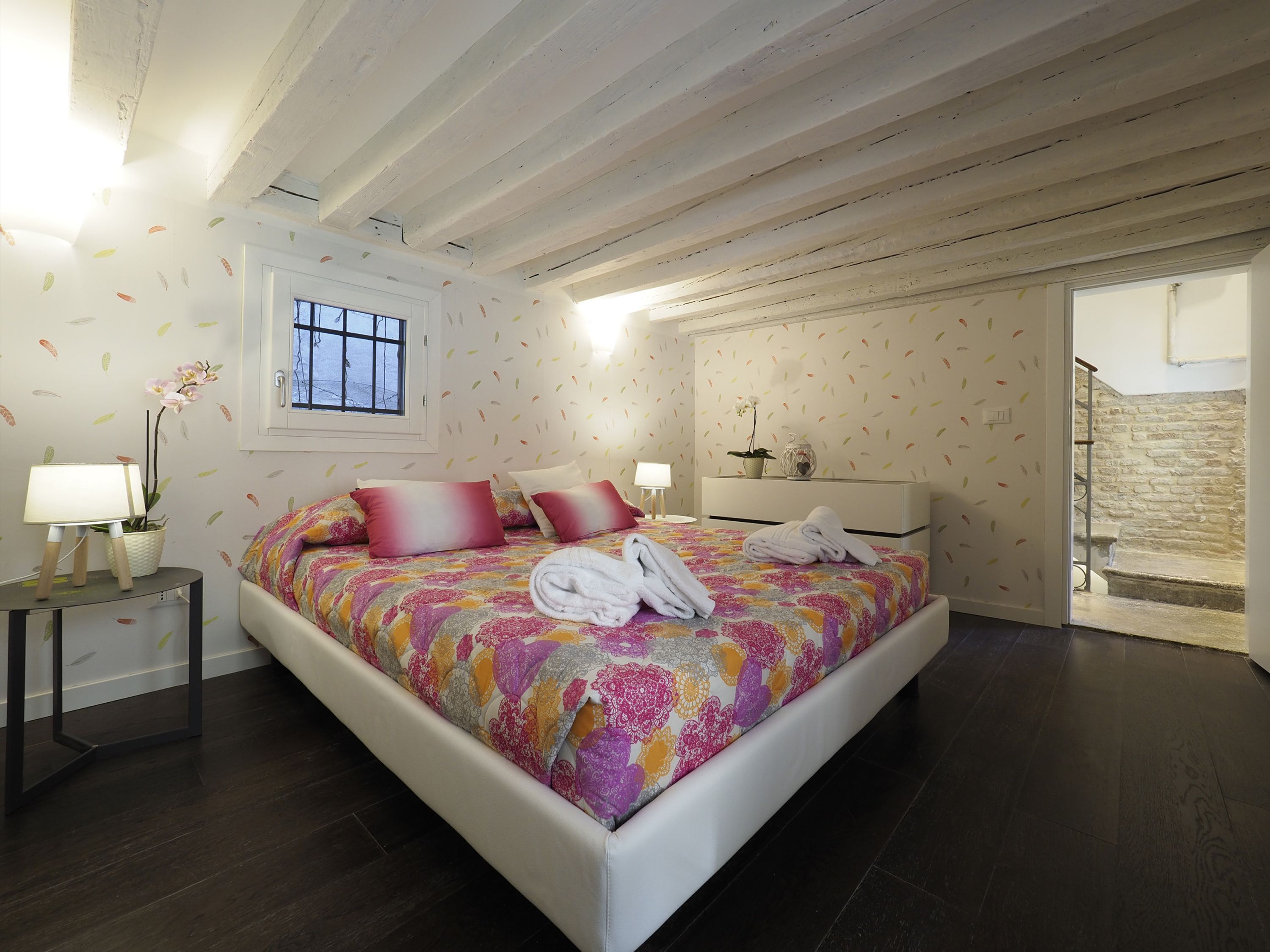 A bedroom with a large bed, a bedside table with a lamp, a window, and a staircase leading to an upper floor.