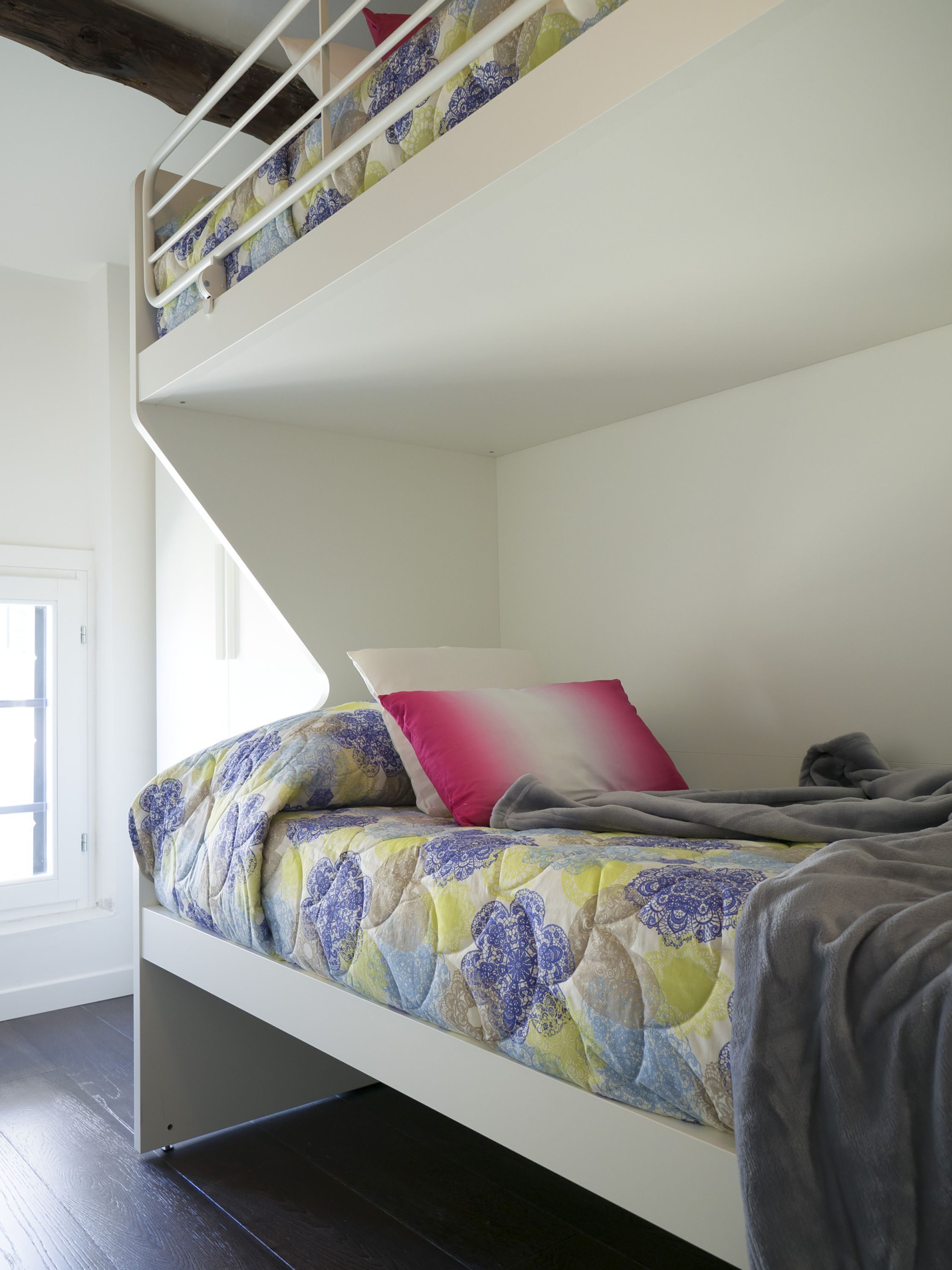A bunk bed with a patterned bedspread and pillows, a white wall, and a window with a view of the outside.