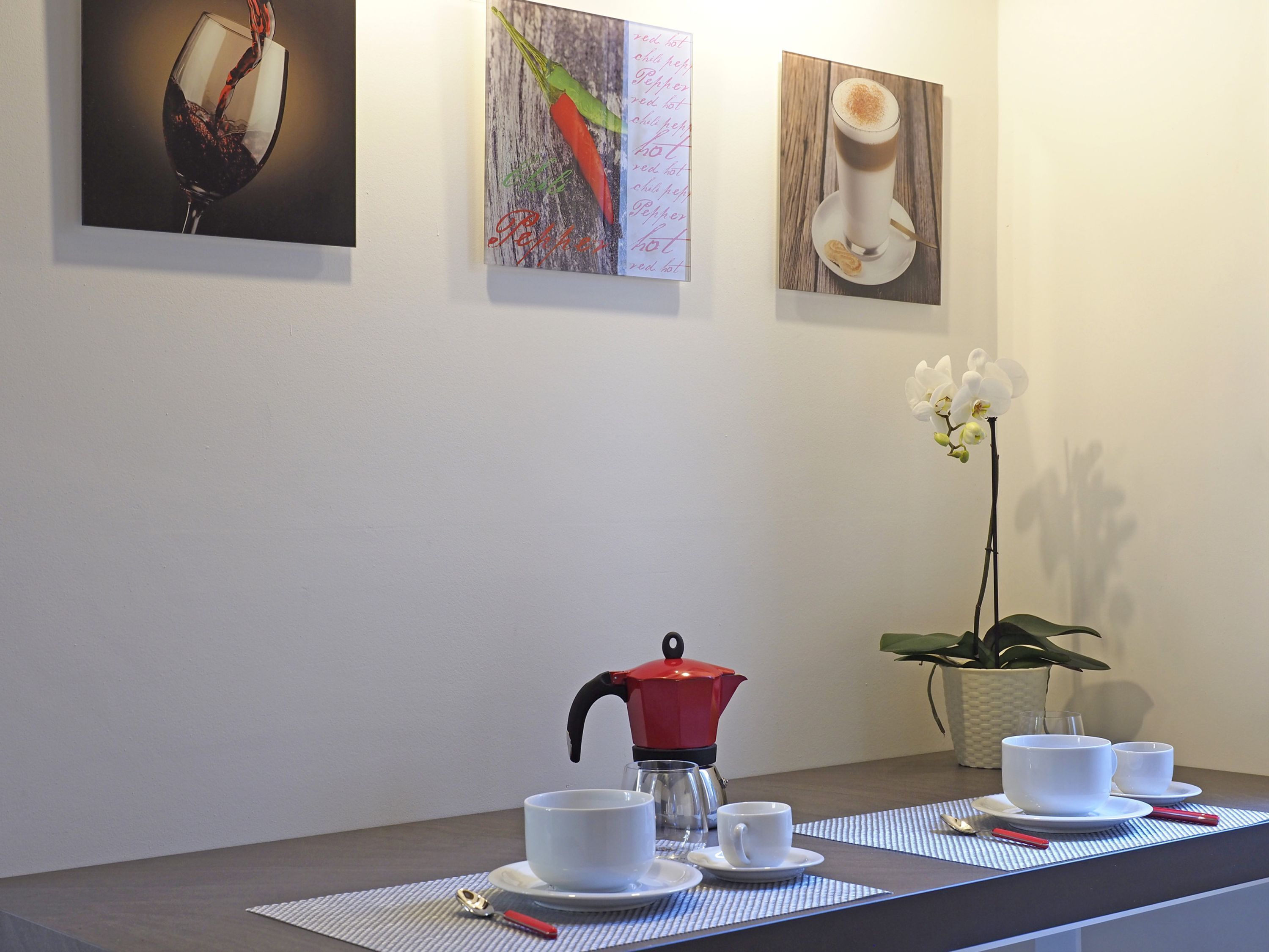 A dining area with a table set for two, a red coffee pot, white cups, and saucers. There are three framed pictures on the wall above the table.