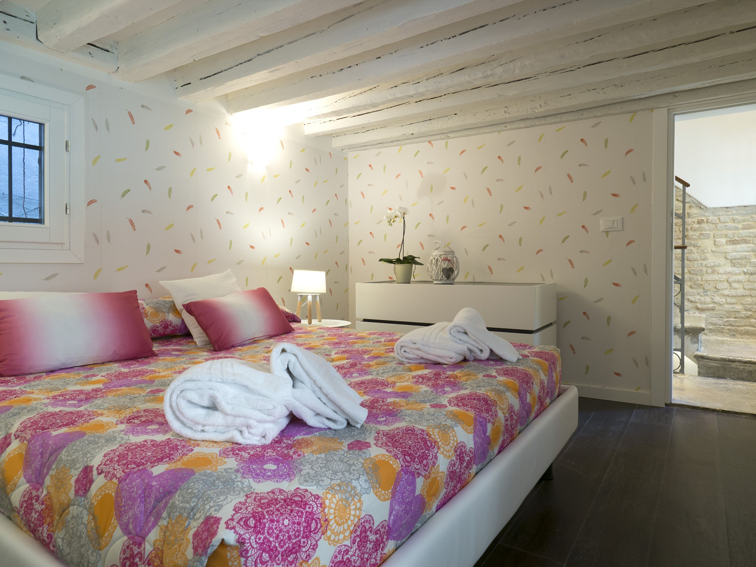 Ein Schlafzimmer mit Bett, Kissen, einem Nachttisch, einer Lampe und Blick ins Freie.