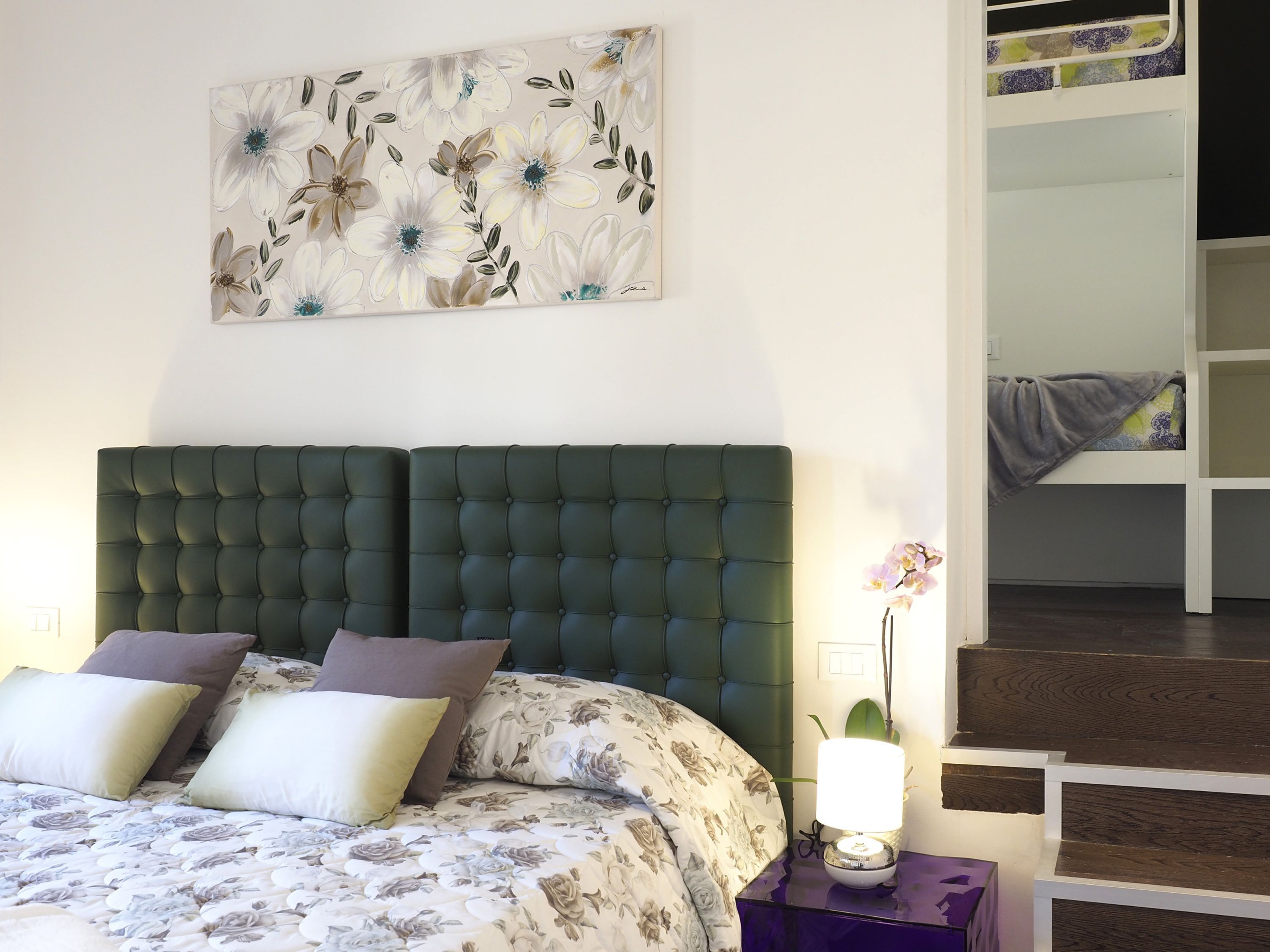 Ein Schlafzimmer mit einer gepolsterten Kopfteile, floralem Bettwäsche, einer Nachttischlampe und einer Treppe, die zu einer oberen Ebene führt.