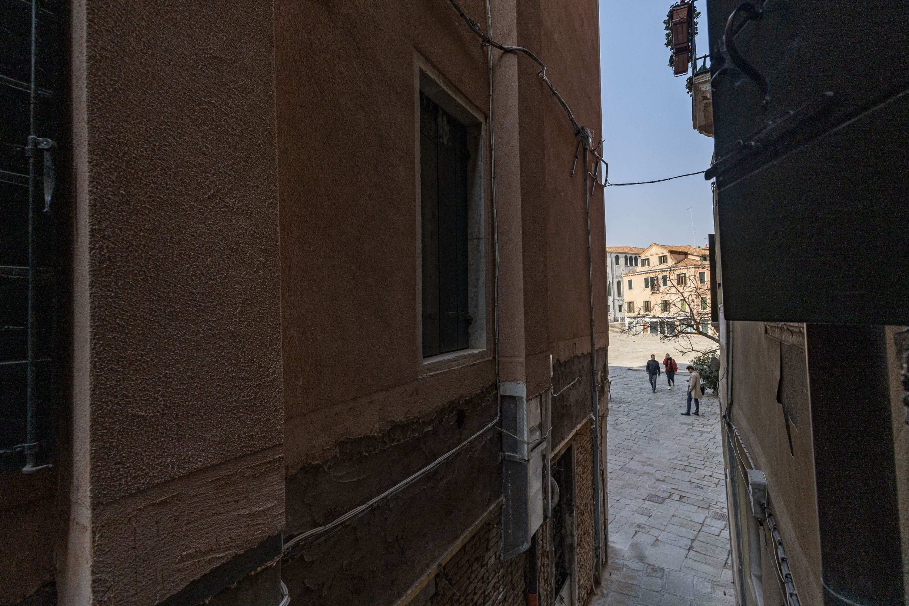 Una calle estrecha flanqueada por edificios, con una ventana visible a la izquierda y algunas personas caminando al fondo.