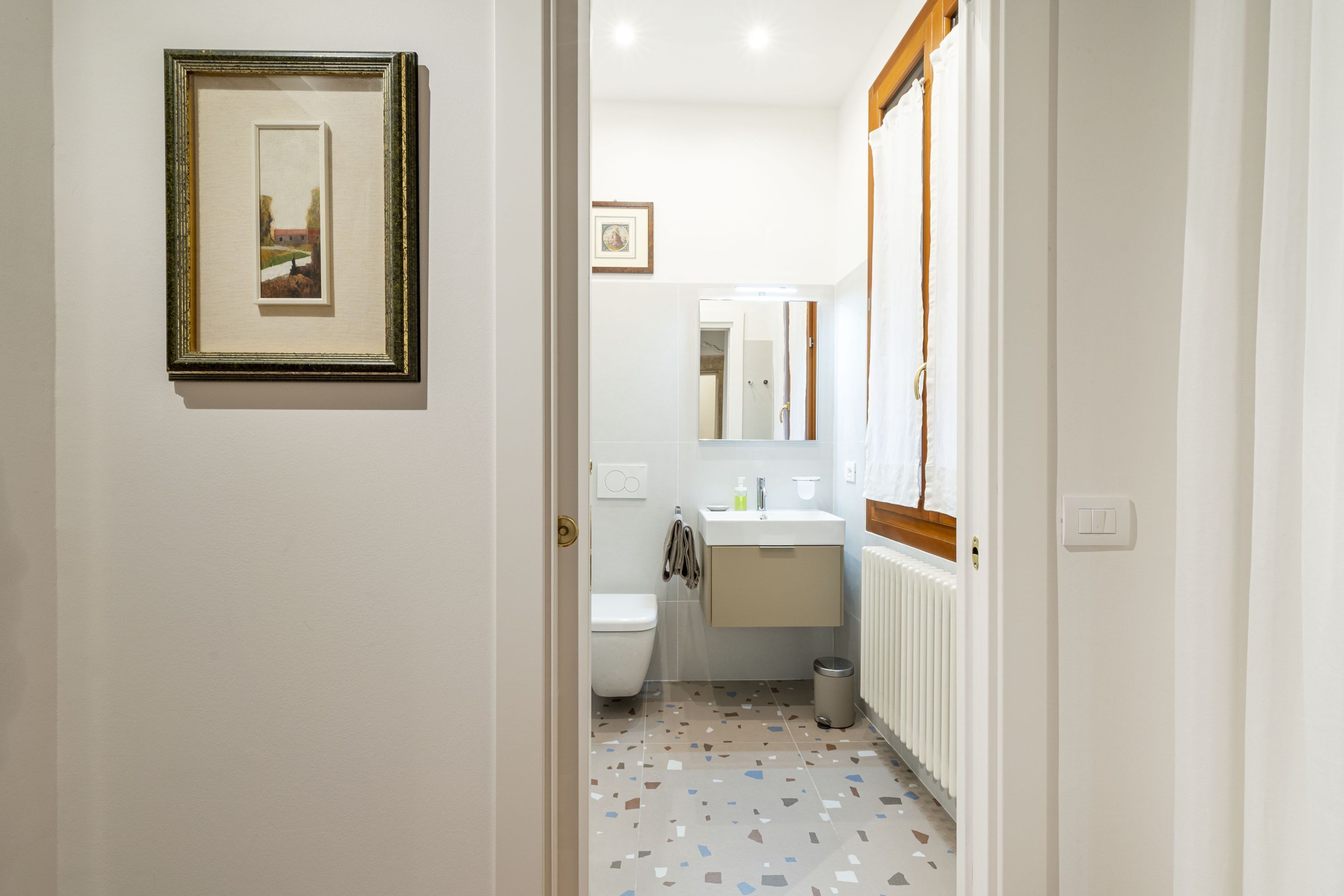 A narrow hallway leading to a bathroom with a toilet, sink, and mirror. A framed picture hangs on the wall.