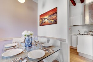 En spiseplads med et bord dækket til fire personer, en vase med blomster og et maleri af en by på væggen.