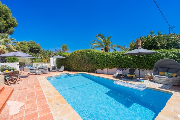 A swimming pool with tiled flooring, surrounded by lounge chairs and umbrellas, and a hedge in the background.
