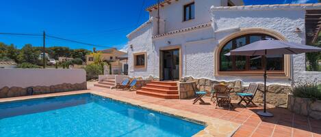 Una casa de dos pisos con piscina, mobiliario exterior y un cielo azul despejado.