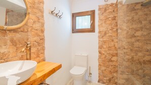Een badkamer met een stenen muur, een houten wastafelmeubel, een wit wastafel, een toilet en een douchehoek.