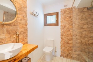 Een badkamer met een stenen muur, een houten wastafelmeubel, een wit wastafel, een toilet en een douchehoek.