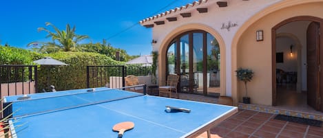 An outdoor ping pong table with a paddle, a patio area with chairs, and a building with arched doorways.