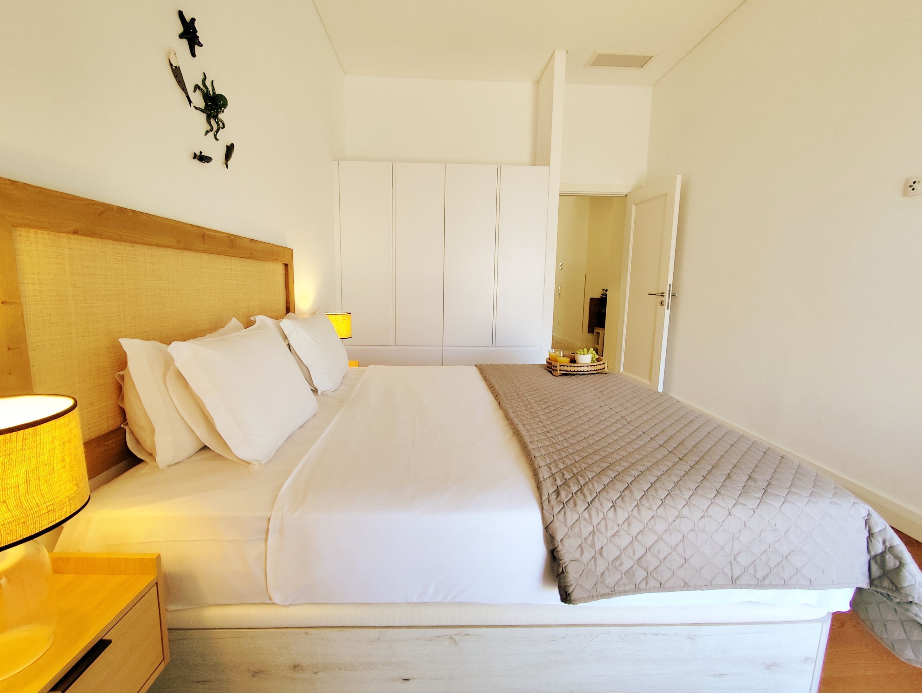 A neatly made bed with white linens and a quilted throw blanket. There is a wooden headboard, a bedside table with a lamp, and a small basket with fruits.